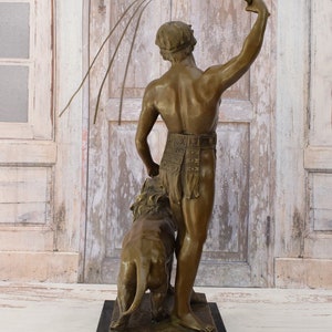 May include: Bronze sculpture of a man holding a whip and standing next to a lion. The sculpture is on a black base.