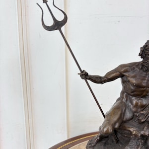 May include: Bronze statue of a muscular male figure holding a trident. The statue is detailed with a dark brown patina, depicting a mythological or historical figure. The trident is held high, and the figure is seated on a rocky base.