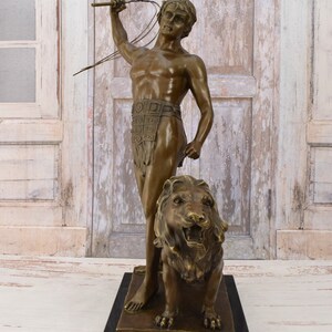 May include: Bronze sculpture of a man holding a whip standing next to a lion. The man is wearing a loincloth and a belt with decorative squares. The lion is roaring with its mouth open. The sculpture is on a black base.