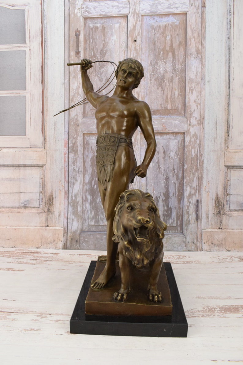 May include: Bronze sculpture of a man holding a whip standing next to a lion. The man is wearing a loincloth and a belt with decorative squares. The lion is roaring with its mouth open. The sculpture is on a black base.