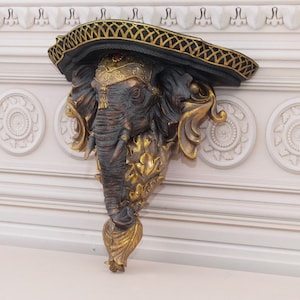 May include: An ornate elephant head wall shelf, featuring a black and gold hat. The elephant is dark grey with gold detailing. The shelf is mounted on a white wall with decorative panelling.