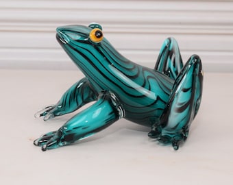 May include: A teal blue glass frog figurine with black stripes and yellow eyes. The frog is sitting on a white surface.