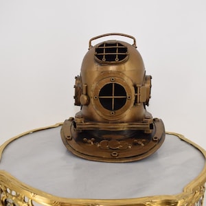 Può includere: Un casco da sub in ottone vintage con una piastra frontale rotonda e oblò multipli. Il casco ha una maniglia sulla parte superiore ed è esposto su un tavolo con piano in marmo e una cornice dorata ornata. È un oggetto decorativo.