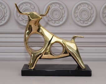 Puede incluir: Una escultura abstracta de toro de color dorado con una base de mármol negro. El toro tiene un diseño estilizado con círculos recortados.