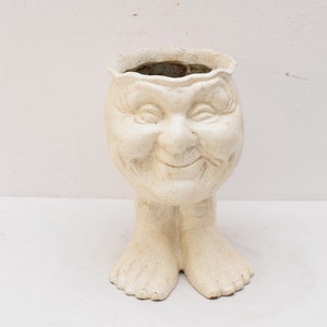 Puede incluir: Maceta de cerámica blanca con forma de cara sonriente con piernas. La maceta tiene una abertura ancha en la parte superior y está diseñada para contener plantas.