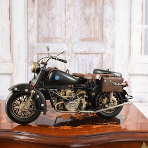 May include: A detailed black and brown vintage motorcycle model with gold accents. The model features a brown leather seat and saddlebags. The motorcycle is displayed on a wooden surface with a decorative edge.