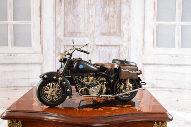 May include: A detailed black and brown vintage motorcycle model with gold accents. The model features a brown leather seat and saddlebags. The motorcycle is displayed on a wooden surface with a decorative edge.