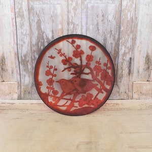 Pode incluir: Um prato redondo de vidro vermelho com um desenho de dois pássaros empoleirados em um galho com flores em flor. O prato tem um efeito de vidro fosco, o que lhe confere uma aparência translúcida.