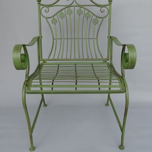 May include: A green metal garden chair with a decorative back featuring fleur-de-lis accents. The chair has curved armrests and a slatted seat. The overall design is ornate and vintage, with a distressed finish.