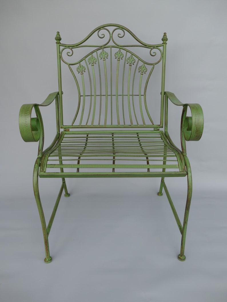 May include: A green metal garden chair with a decorative back featuring fleur-de-lis accents. The chair has curved armrests and a slatted seat. The overall design is ornate and vintage, with a distressed finish.