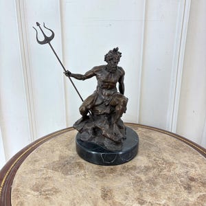 May include: Bronze sculpture of a seated figure holding a trident. The figure is on a rocky base atop a black marble plinth. The sculpture has a dark brown patina and is likely a depiction of a mythological figure.
