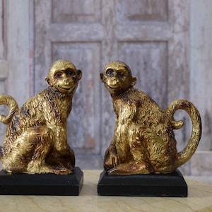 May include: Two gold-colored monkey figurines, each sitting on a black rectangular base. The monkeys have a textured, detailed finish and are posed with a smiling expression. The background is a blurred wooden door.