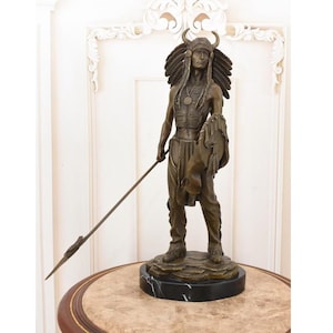 Op de afbeelding: Een bronzen beeld van een Indiaanse stamhoofd, staande met een speer en schild. De figuur draagt een gevederde hoofdtooi en traditionele kleding. Het beeld is gemonteerd op een zwarte marmeren voet, met ingewikkelde details en vakmanschap.