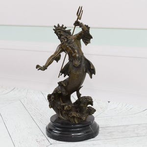 Puede incluir: Estatua de bronce de una criatura marina m&iacute;tica, posiblemente Poseid&oacute;n o Neptuno, sosteniendo un tridente. La escultura presenta intrincados detalles de la cara, la corona y la cola de pez de la figura. La estatua est&aacute; montada sobre una base de m&aacute;rmol negro.