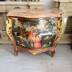 May include: A bombe chest featuring a pink marble top and three drawers. The front displays a painted scene of two figures in a landscape. The piece is decorated with ornate gold-toned metal mounts on the corners, legs, and drawer pulls.