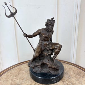 May include: Bronze sculpture of a seated figure holding a trident. The figure has a beard and is atop a rocky base, mounted on a black marble plinth. The sculpture is a dark bronze color.