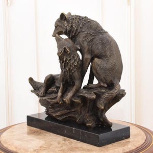 May include: A bronze sculpture of two wolves. One wolf is seated, looking up, while the other stands over it. The sculpture is mounted on a black marble base and sits on a decorative table.