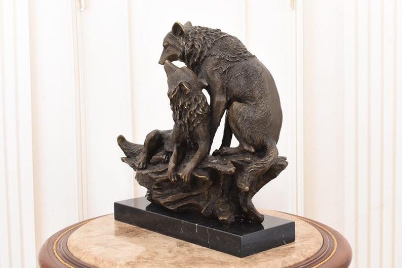 May include: A bronze sculpture of two wolves. One wolf is seated, looking up, while the other stands over it. The sculpture is mounted on a black marble base and sits on a decorative table.
