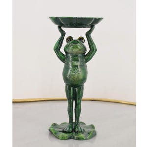 Könnte beinhalten: Eine grüne Froschfigur, die eine gewellte Schale über dem Kopf hält. Der Frosch hat große Augen und steht auf einem Seerosenblatt-Sockel. Die Figur ist ein dekoratives Element.