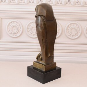 May include: A bronze sculpture of an owl with a black marble base. The owl is standing on a base and is facing to the right. The owl is stylized and has a geometric design.