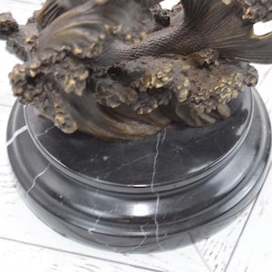 Puede incluir: Una escultura de bronce de una sirena emergiendo de las olas del oc&eacute;ano. La escultura est&aacute; montada sobre una base redonda de m&aacute;rmol negro con vetas blancas. La cola de la sirena y las olas est&aacute;n detalladas.