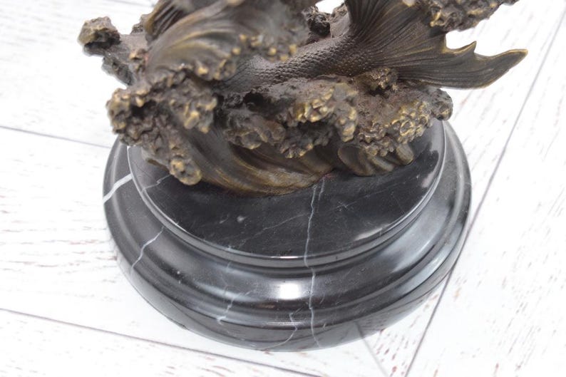 Puede incluir: Una escultura de bronce de una sirena emergiendo de las olas del oc&eacute;ano. La escultura est&aacute; montada sobre una base redonda de m&aacute;rmol negro con vetas blancas. La cola de la sirena y las olas est&aacute;n detalladas.