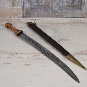 May include: A double-edged short sword with a wooden handle and brass accents, featuring a central fuller along the blade. Beside the sword lies a black scabbard with a brass tip and a brass fitting at the top, designed for storage and protection.