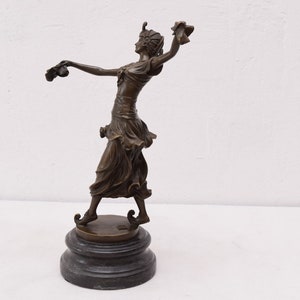 Op de afbeelding: Bronzen beeld van een vrouw in een vloeiende jurk, met in elke hand een bloem, staand op een ronde basis.