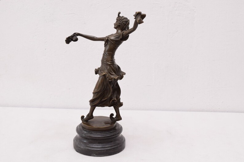 Op de afbeelding: Bronzen beeld van een vrouw in een vloeiende jurk, met in elke hand een bloem, staand op een ronde basis.