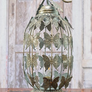 Può includere: Una lampada a lanterna in metallo decorativa con una struttura a gabbia ovale ornata da numerosi ritagli a forma di farfalla. La lampada presenta una finitura anticata color oro e verde, una base circolare e un manico superiore curvo.