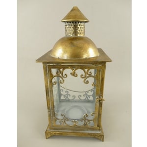 Può includere: Una lanterna in metallo color oro con una parte superiore a cupola e un piccolo tetto a punta. La lanterna ha pannelli di vetro trasparente e volute ornate sulla parte anteriore e sui lati. C'è una piccola porta sul lato e un portacandele all'interno.