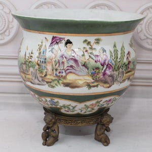 Puede incluir: Un macetero de porcelana blanca con un borde verde y diseños florales. El macetero presenta una escena de personas en un entorno de jardín. El macetero se asienta sobre un soporte de metal dorado con tres patas.