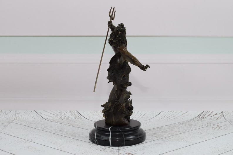 Puede incluir: Escultura de bronce de una figura sosteniendo un tridente, montada sobre una base de m&aacute;rmol negro. La figura tiene una apariencia detallada y texturizada, con ropas fluidas y una corona. La escultura se encuentra sobre un fondo blanco y celeste.