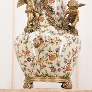 May include: A decorative ceramic vase with a floral pattern in orange, blue, and purple on a cream background. The vase features bronze cherub figurines and a decorative base. The vase is a classic home decor item.