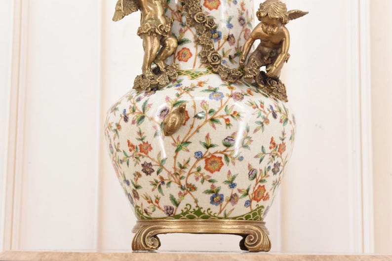 May include: A decorative ceramic vase with a floral pattern in orange, blue, and purple on a cream background. The vase features bronze cherub figurines and a decorative base. The vase is a classic home decor item.