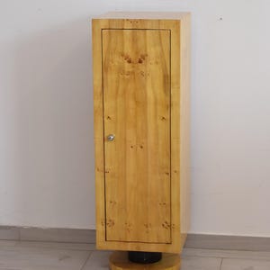 Puede incluir: Armario alto de madera clara con una sola puerta y un pequeño tirador redondo plateado. El armario tiene forma rectangular y se apoya sobre una base circular. Se aprecia la veta de la madera.