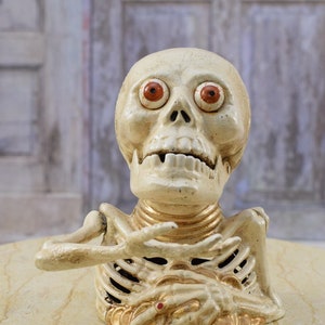 Puede incluir: Una alcancía de cerámica blanca en forma de esqueleto con ojos rojos y monedas de oro en su regazo. El esqueleto está sentado sobre una superficie beige claro.