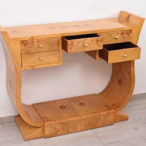 May include: A light-colored wooden console table with a curved base and top. It features five drawers with silver and gold hardware. The drawers are open, revealing the interior. The table is made of wood with a unique grain pattern.