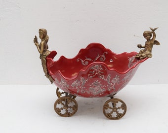 May include: A red ceramic bowl with a white floral design, mounted on a wheeled base with gold cherub figures on each side.