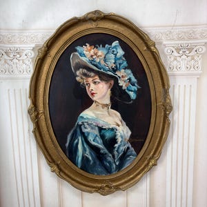 Op de afbeelding: Een ovaal olieverfschilderij in een decoratieve gouden lijst met een persoon in een blauwe jurk en een bijpassende blauwe hoed versierd met bloemen. Het onderwerp is vanaf de borst afgebeeld en kijkt naar links tegen een donkere achtergrond.