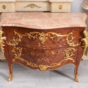 May include: A brown antique chest of drawers with a pink marble top and gold accents. The chest has two drawers and ornate gold detailing, including floral and figure motifs. The legs are also gold.