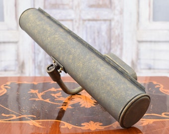 May include: A brass picture light with a long, cylindrical shade. The shade has a distressed finish and is angled downwards. The light is mounted on a wall.