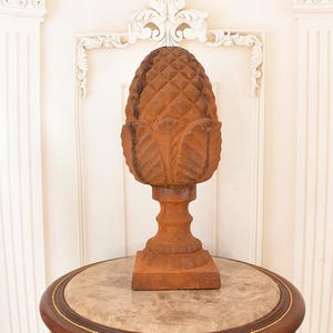 May include: A large, weathered, brown cast iron pineapple sculpture. The pineapple sits atop a pedestal base and is displayed on a round table with a marble top. The sculpture is a decorative accent piece.