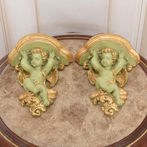 May include: Two decorative wall shelves featuring cherub figures. Each shelf has a light green top with gold trim, supported by a cherub statue in a seated position. The cherubs are light green with gold accents, and the shelves are placed on a round table.