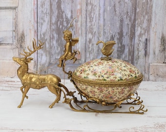 May include: A gold-tone reindeer pulls a sleigh with a porcelain egg-shaped box. The box is decorated with a floral pattern and topped with a swan figurine. The reindeer is detailed with a harness and a rider.