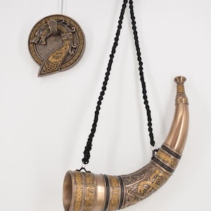 Peut inclure: Une corne à boire de couleur bronze avec des entrelacs celtiques complexes et une corde noire pour la suspendre. Une plaque murale ronde assortie avec un motif de dragon est également visible. La corne mesure environ 30 cm de long.