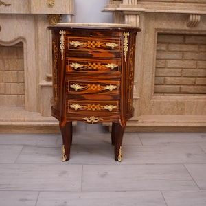 May include: A small, antique wooden cabinet with four drawers. The cabinet is a rich brown with a striped pattern and gold-colored accents. The drawers have gold handles and floral designs. The cabinet has four legs and is placed on a light gray floor.