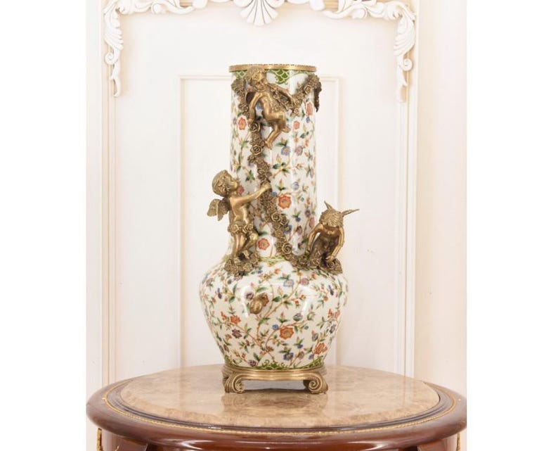 May include: A tall, ornate ceramic vase with a floral pattern in shades of red, orange, and blue. The vase features bronze cherub and owl embellishments, and sits on a decorative base. The vase is approximately 24 inches tall.