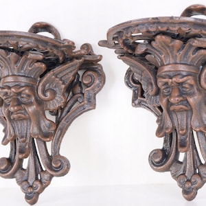 Puede incluir: Dos estanterías de pared ornamentadas de hierro fundido con acabado bronce. Cada estantería presenta un rostro detallado con barba, alas y adornos. Tienen una parte superior pequeña y curvada y un lazo para colgar.