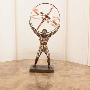 Puede incluir: Estatua de bronce de Atlas sosteniendo una esfera armilar de color cobre. La escultura está sobre una base rectangular y sobre una superficie con vetas de mármol. Un artículo decorativo.